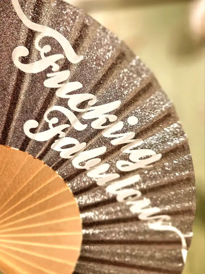 Dekorativ sølv vifte med "Fucking Fabulous" tekst i glitter, perfekt til fest og arrangementer.